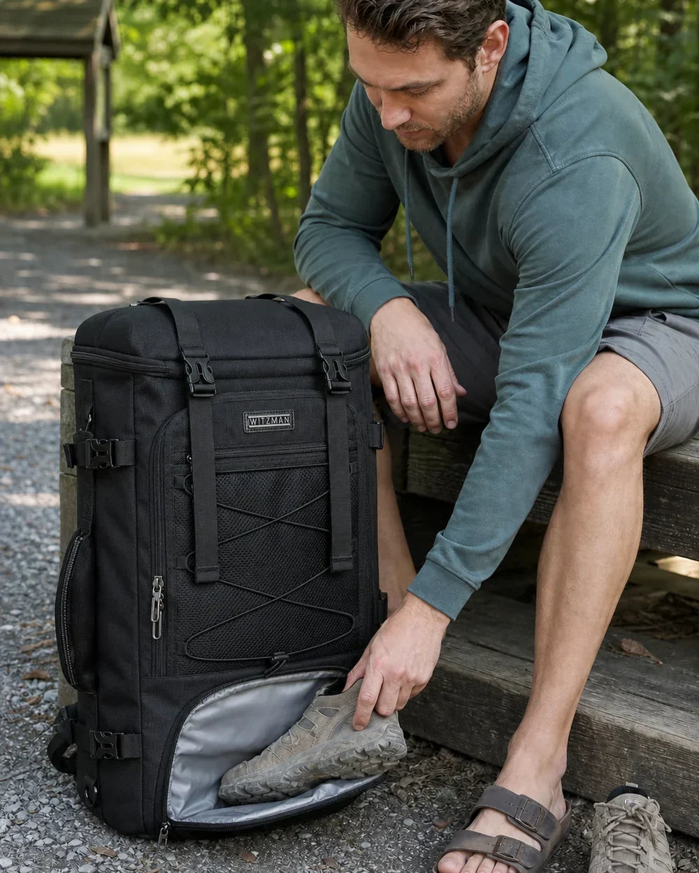 Traveler placing dusty trail shoes into the lower compartment of a full-size black WITZMAN B715 travel backpack at a wooded trailhead
